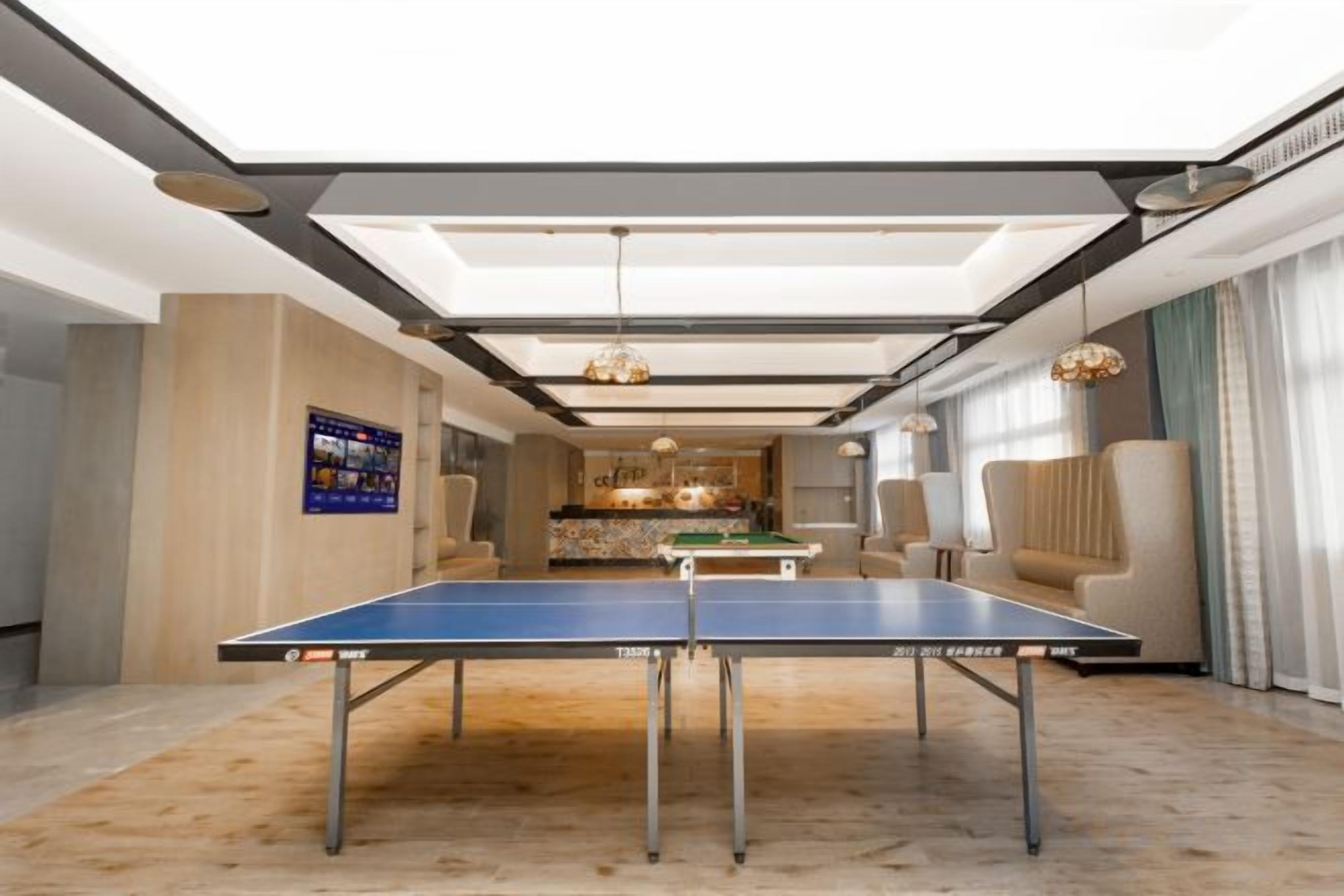 Ett bordtennisbord i ett modernt rum med högt i tak, en matplats i bakgrunden och utsikt över utomhusmiljön genom stora fönster.