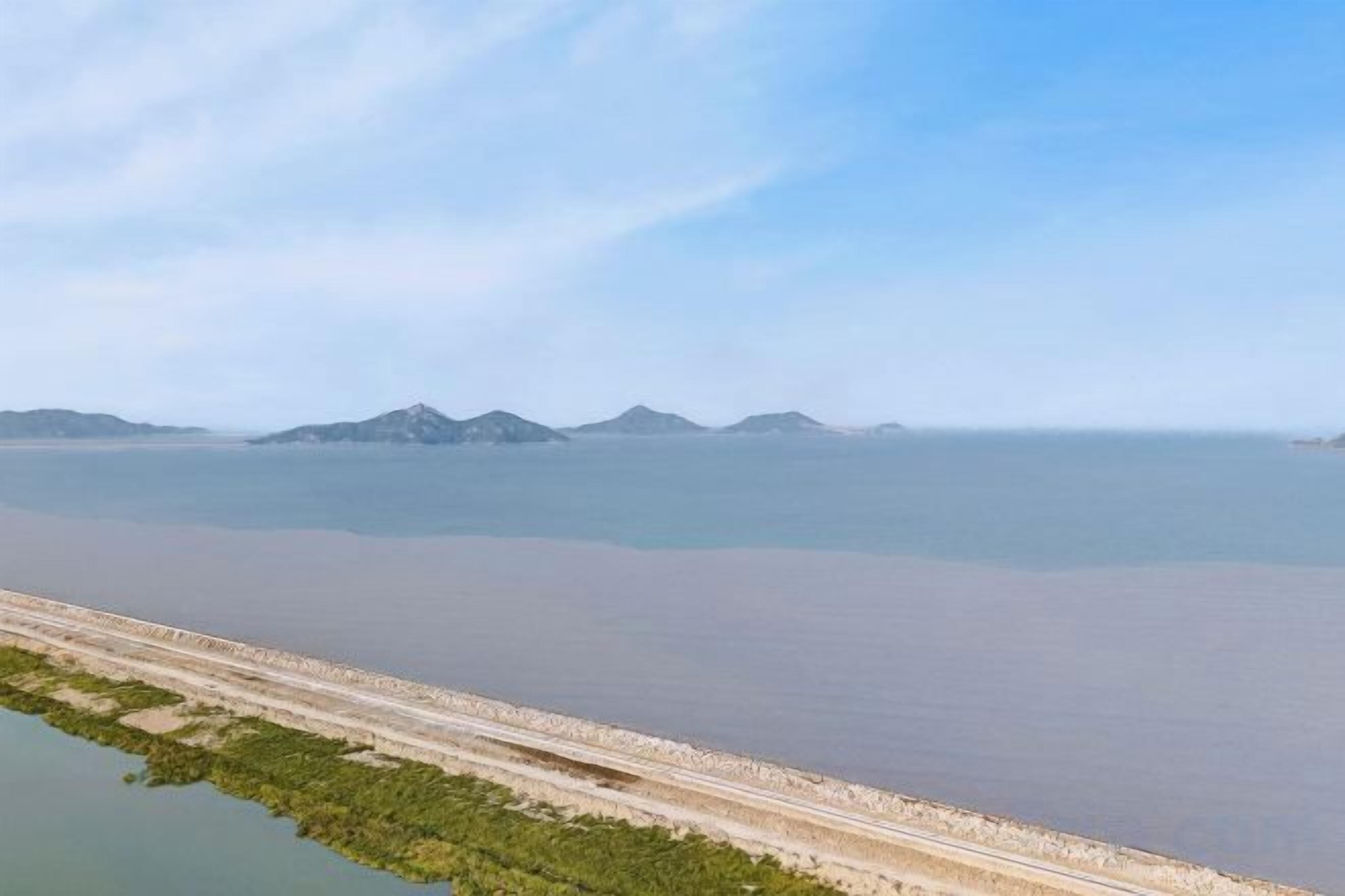A coastal landscape with a long, narrow strip of land leading to the sea, a clear sky, and distant mountains.