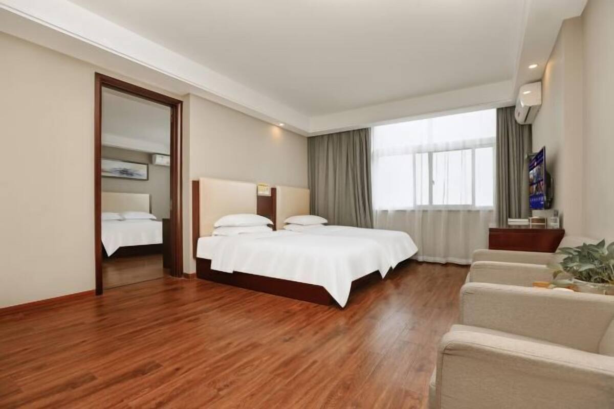 Habitación de hotel con suelo de madera, cama doble, zona de estar con sofá y ventana grande con cortinas.