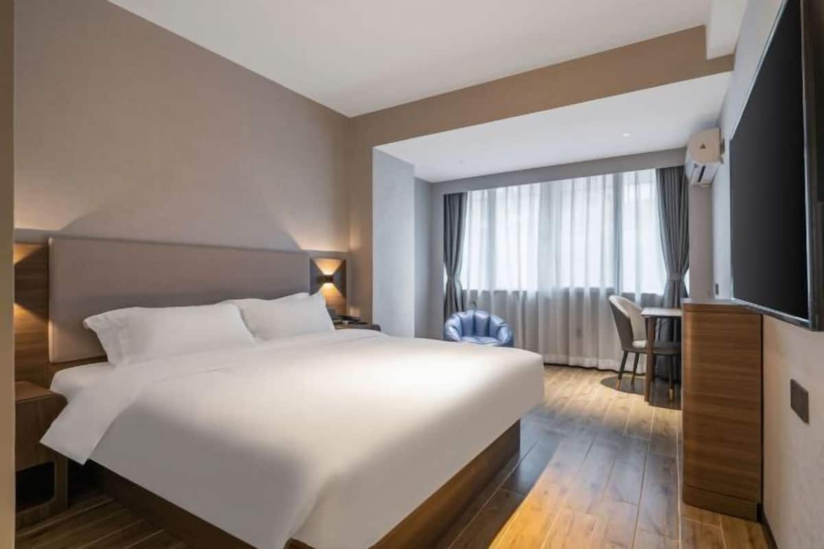 A modern hotel room with a large bed, a desk, and a chair, a flat-screen TV, and a large window with curtains.