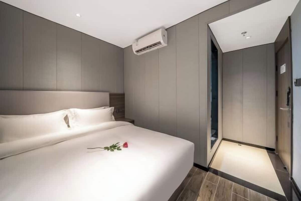 A modern hotel room with a large bed, a bedside table, an air conditioning unit, and a shower area.