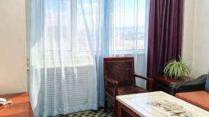 Une chambre d’hôtel avec un sol à carreaux, une chaise en bois, une table avec un magazine, une plante et des rideaux en voile.