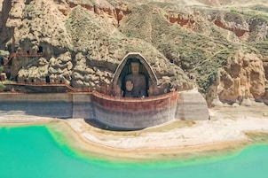 Una estatua grande de Buda dentro de una estructura similar a una cueva, rodeada de un paisaje rocoso y una masa de agua.