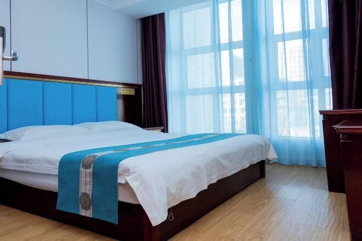 Een hotelkamer met een bed, een houten vloer, een raam met gordijnen en een houten dressoir.