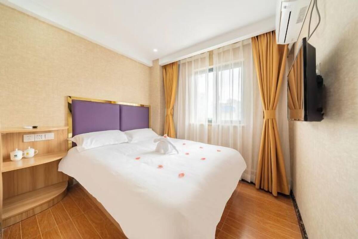 A hotel room with a bed, a bedside table with a lamp, a TV mounted on the wall, and a window with curtains.