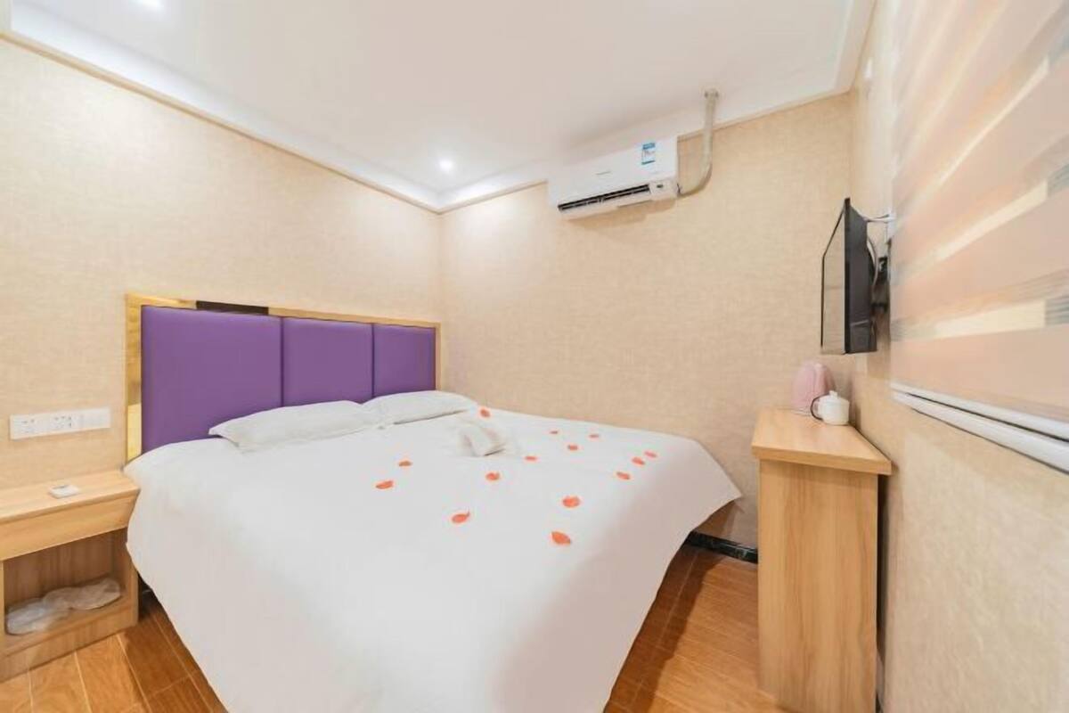 A hotel room with a bed, a nightstand, an air conditioning unit, and a television mounted on the wall.