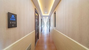 Pasillo de hotel con un panel de número de habitación digital, suelo de madera y un largo pasillo con puertas a ambos lados.