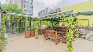 Un patio al aire libre con un banco y una mesa de madera, rodeado de follaje y un suelo de baldosas.