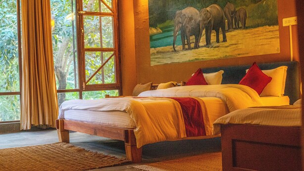 A room with a large bed, a painting of elephants on the wall, and a view of greenery through the window. - Jungle Adventure World Hotel Pvt. Ltd (Sauraha)