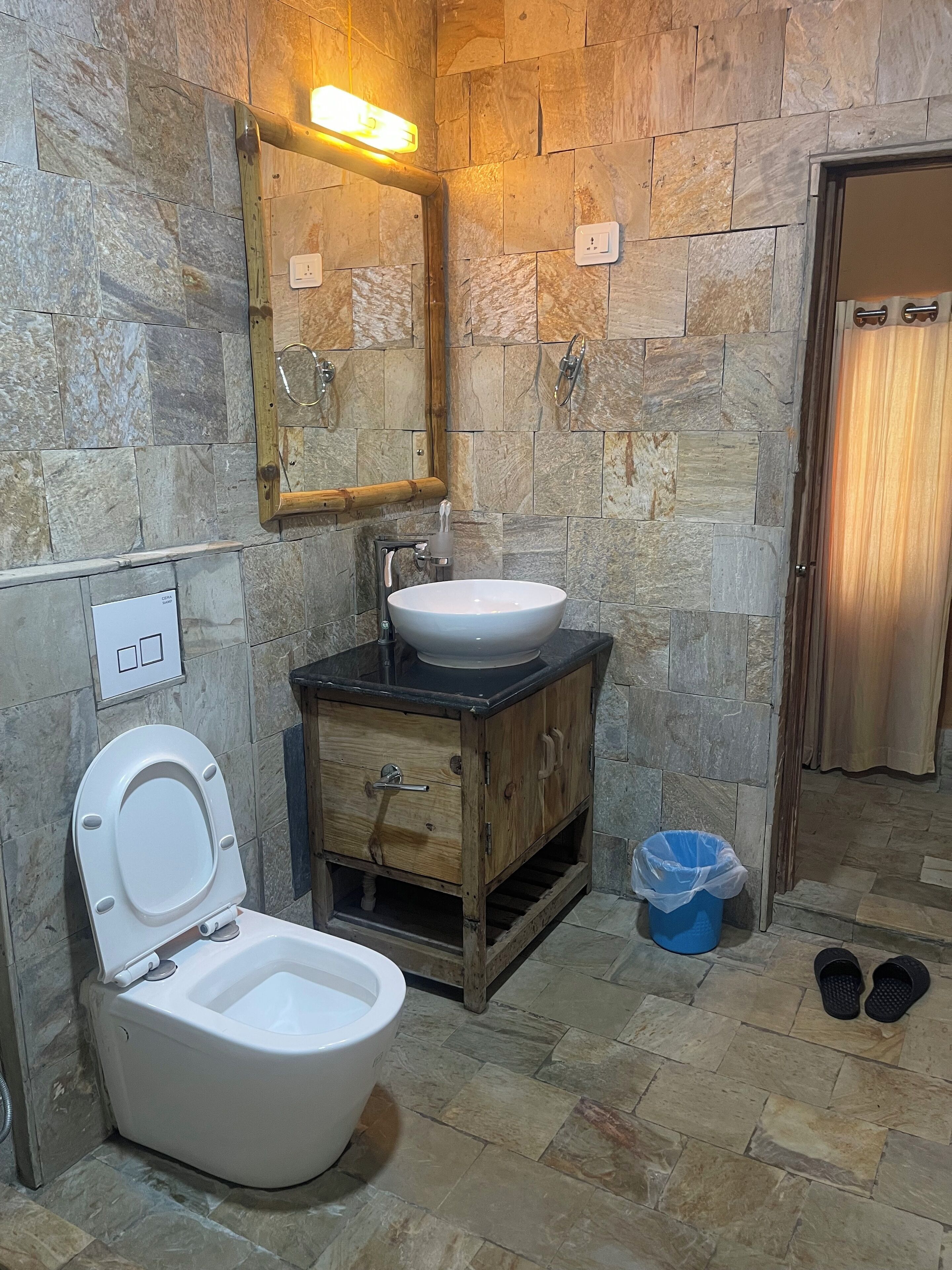 Baño con inodoro, lavamanos, mueble de madera, paredes de piedra, espejo y cortina de ducha.