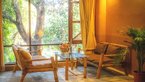A room with a glass table, two wooden chairs, a sofa, a potted plant, and a view of greenery outside. - Jungle Adventure World Hotel Pvt. Ltd (Sauraha)