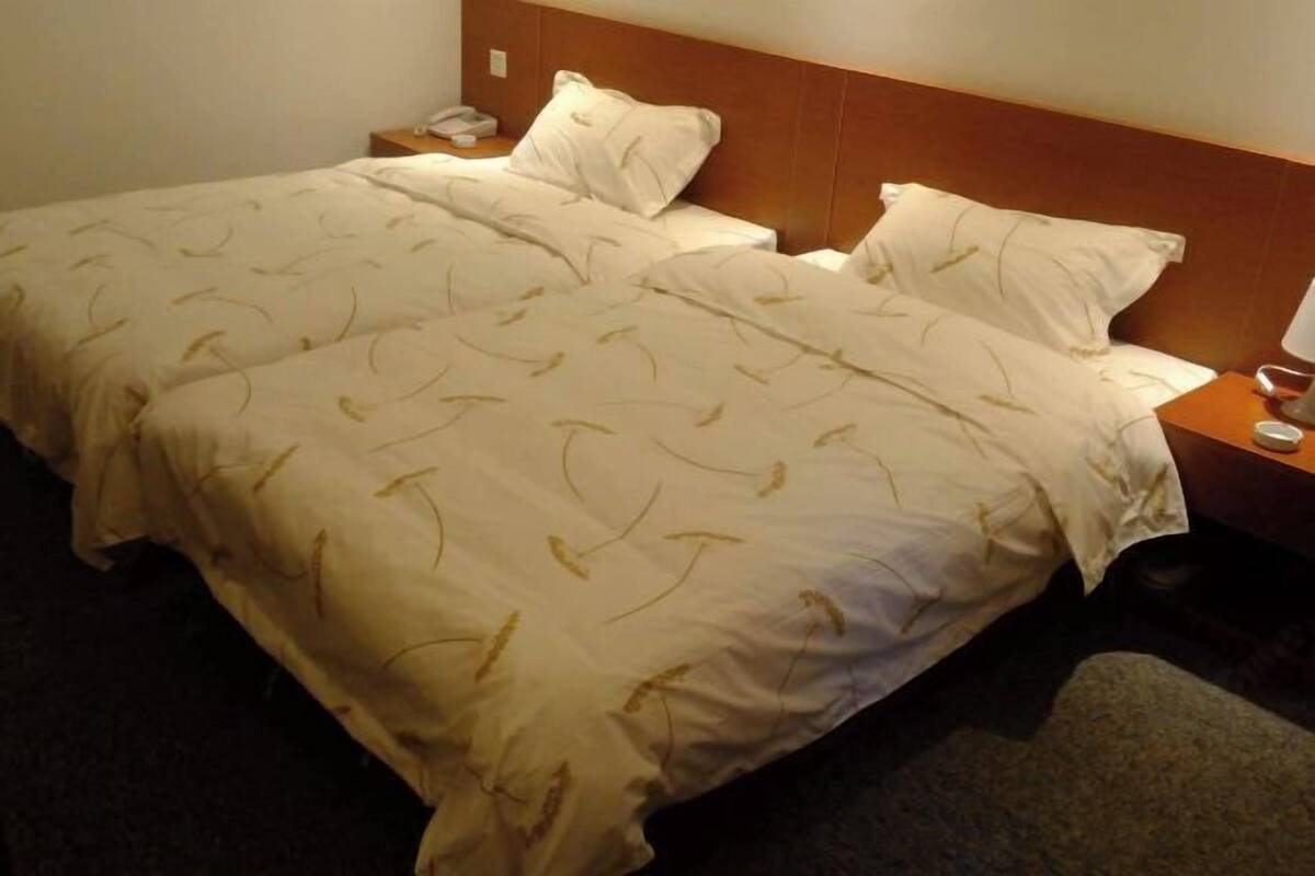 A double bed with a white bedspread featuring a leaf pattern, a wooden headboard, a bedside table with a lamp, and a phone.