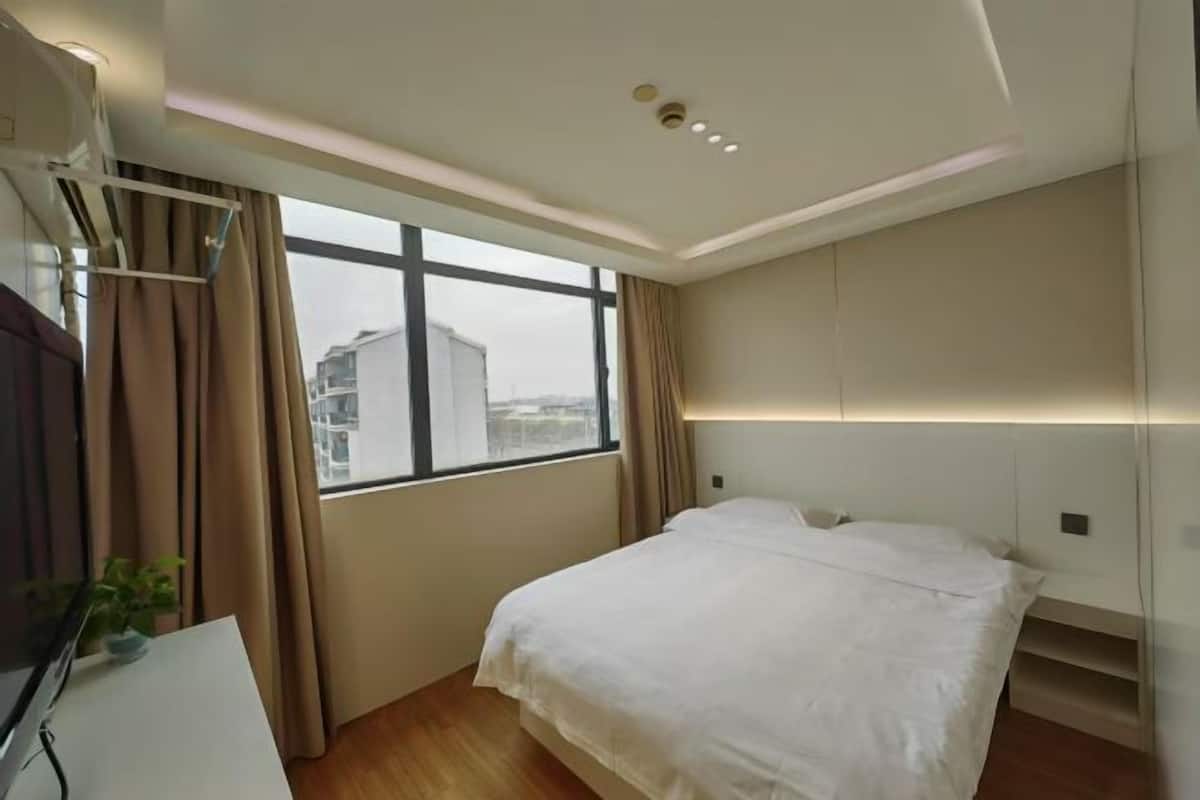 A hotel room with a bed, a desk, a window with curtains, and a view of buildings outside.