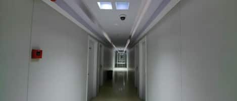 A narrow, well-lit hallway with a ceiling light fixture and a fire alarm on the left wall.