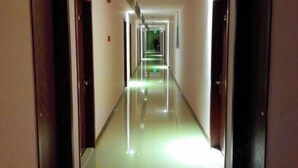 Corredor longo e estreito de um hotel, com portas de ambos os lados e piso brilhante que reflete a luz.