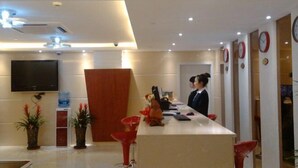 Uma área de recepção de hotel, com balcão de atendimento, televisão fixada na parede e um recepcionista.