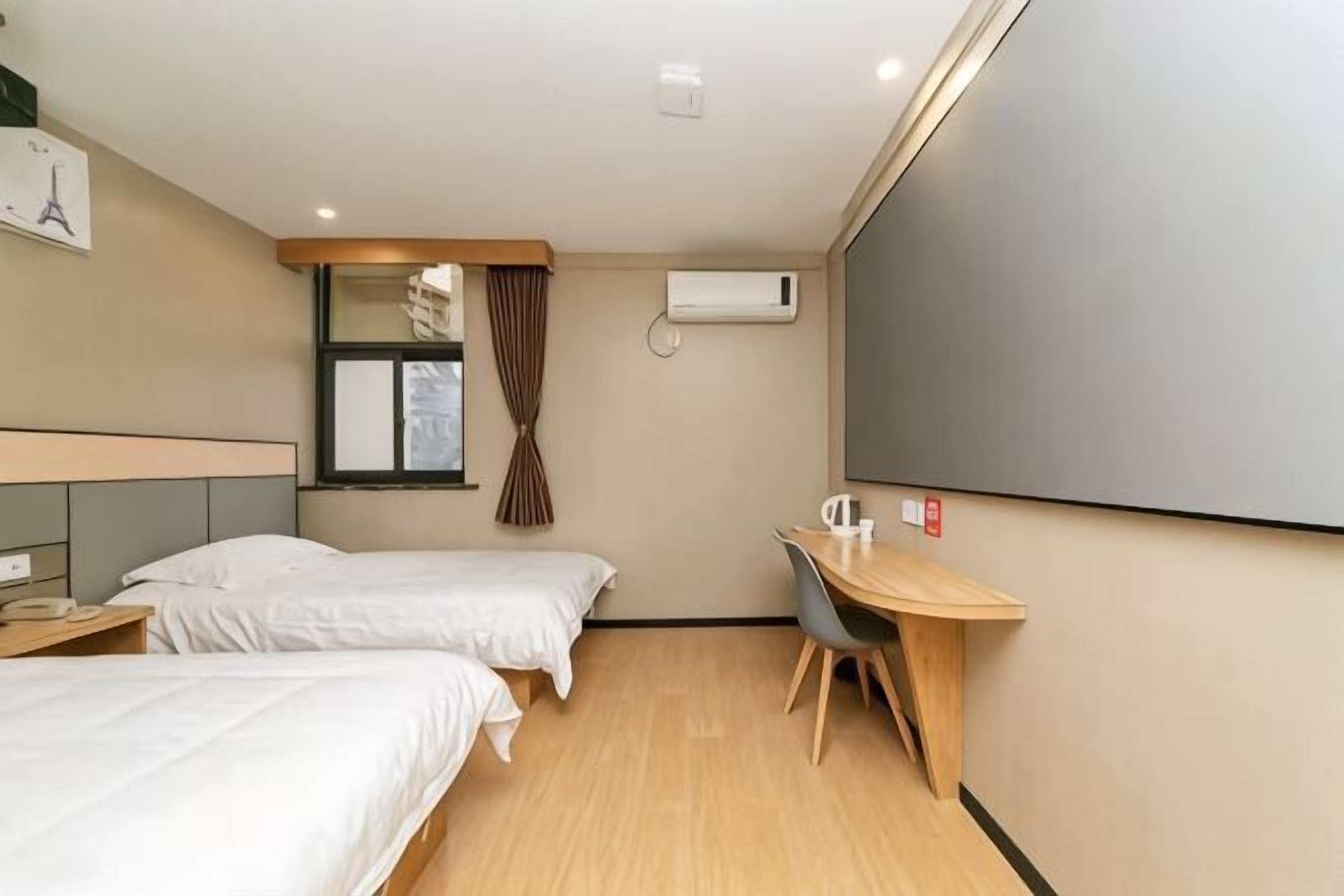 A hotel room with two beds, a desk, a chair, a window with curtains, an air conditioning unit, and a wall-mounted television.