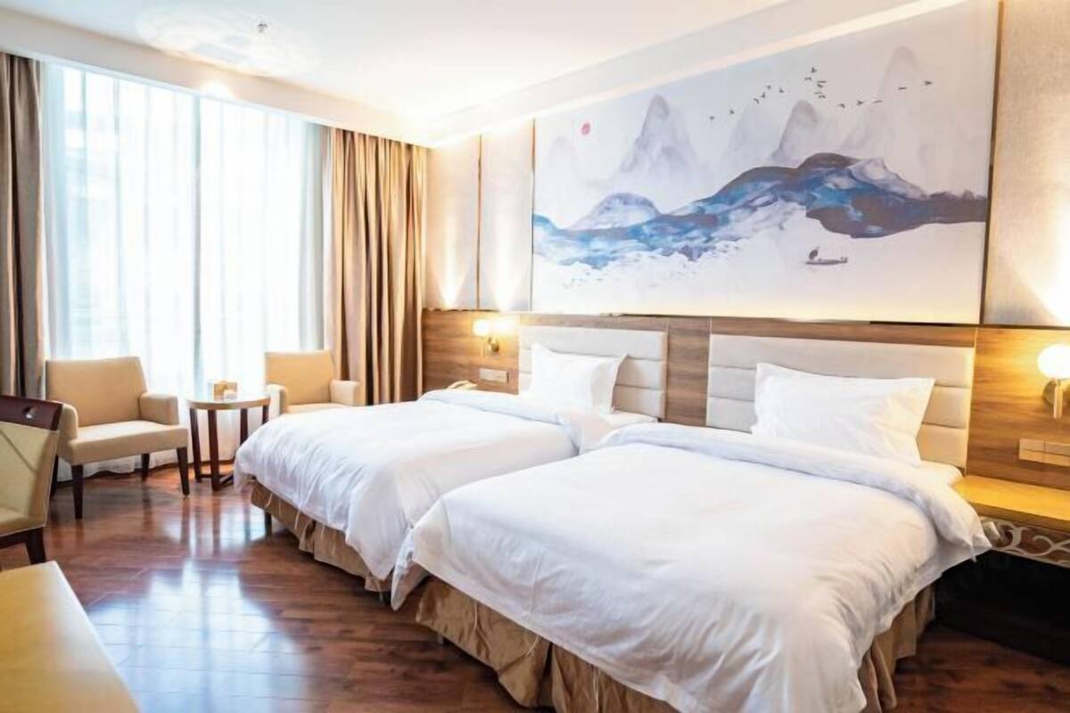 A hotel room with two beds, a sitting area with a chair and a small table, and a large painting of mountains on the wall.