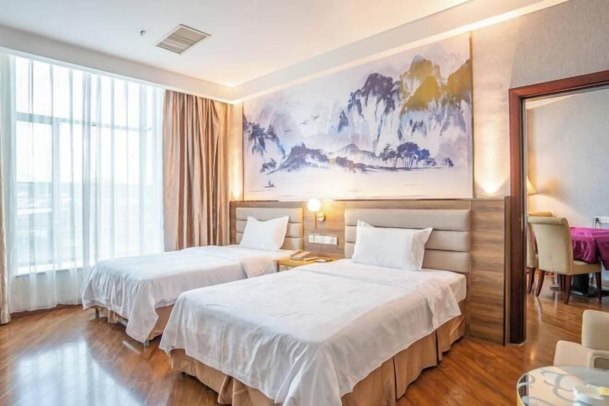 A hotel room with two beds, a large painting on the wall, and a dining area visible through the window.