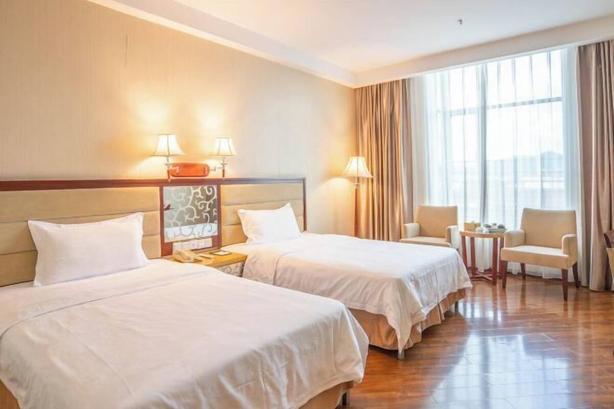 A hotel room with two beds, a small table, a chair, and a large window with curtains.