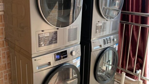 A stacked washer and dryer unit with a digital display, located in a room with a brick wall and a window with red curtains. - Federal Hotel (Meizhou)