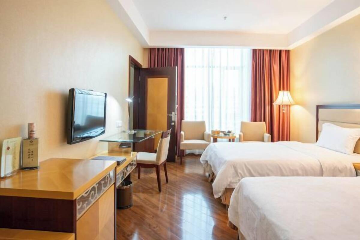 A hotel room with a TV, desk, two beds, a chair, and a window with curtains.