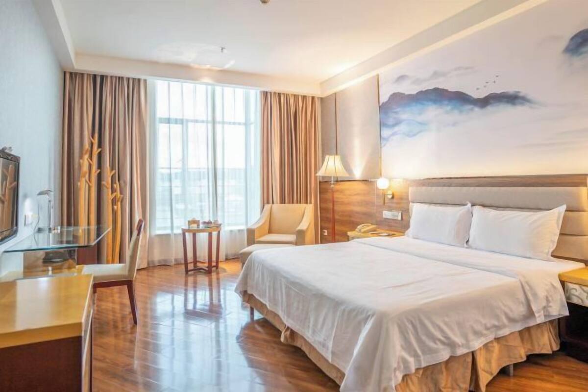 A hotel room with a large bed, a desk with a chair, a small table, and a window with curtains.