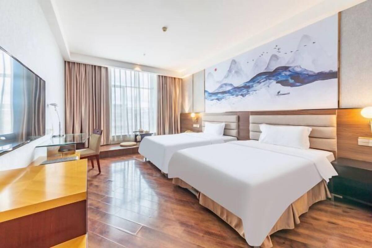 A hotel room with two beds, a desk, a large window with curtains, and a painting on the wall.