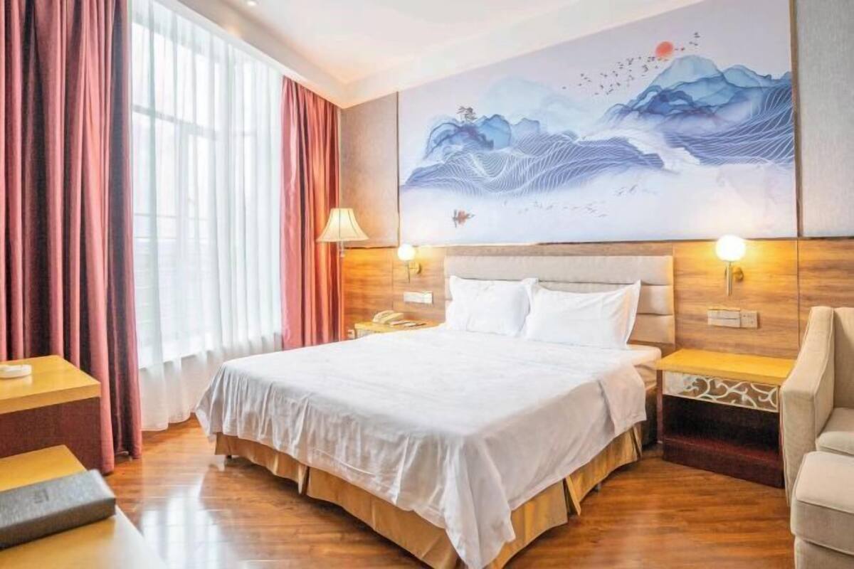 A hotel room with a large bed, wooden headboard, two bedside tables, a nightstand, a lamp, and a painting on the wall.