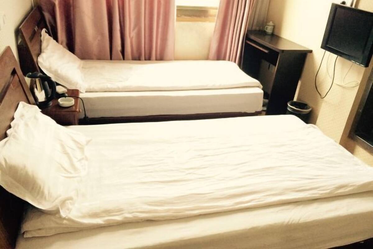 A hotel room with two single beds, a bedside table with a lamp, a TV mounted on the wall, and a window with curtains.
