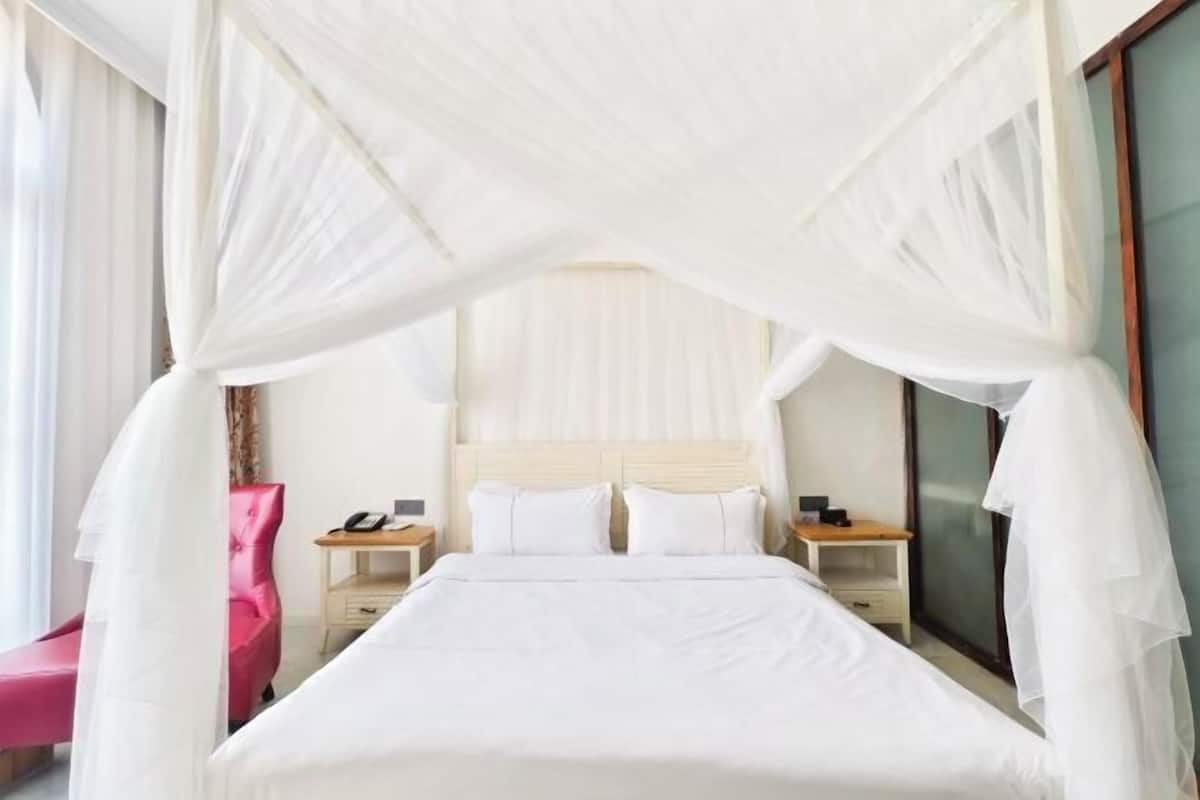 A hotel room with a bed, two bedside tables, a nightstand, a pink chair, and a large window.