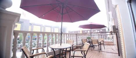 A balcony with wooden chairs and tables, a large umbrella, and a railing.