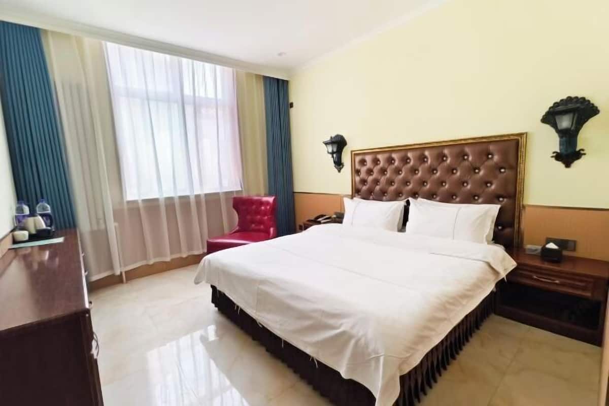 A hotel room with a large bed, a red chair, a bedside table, a lamp, and a window with curtains.