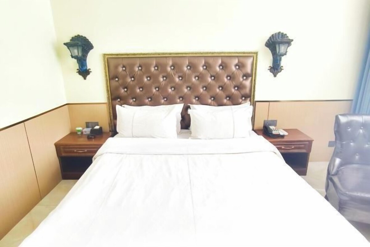 Una habitación de hotel con una cama grande, dos mesitas de noche, una silla y lámparas fijadas a la pared.