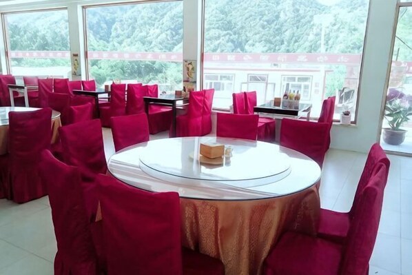 A dining area with round tables and red chairs, large windows with a view of greenery, and a white tablecloth on one table.
