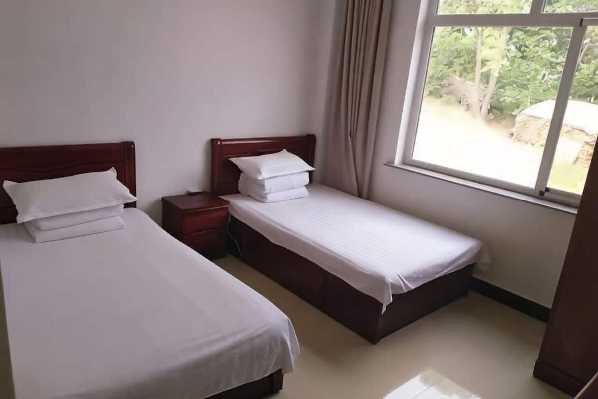 A room with two beds, a window with curtains, and a view of greenery outside.