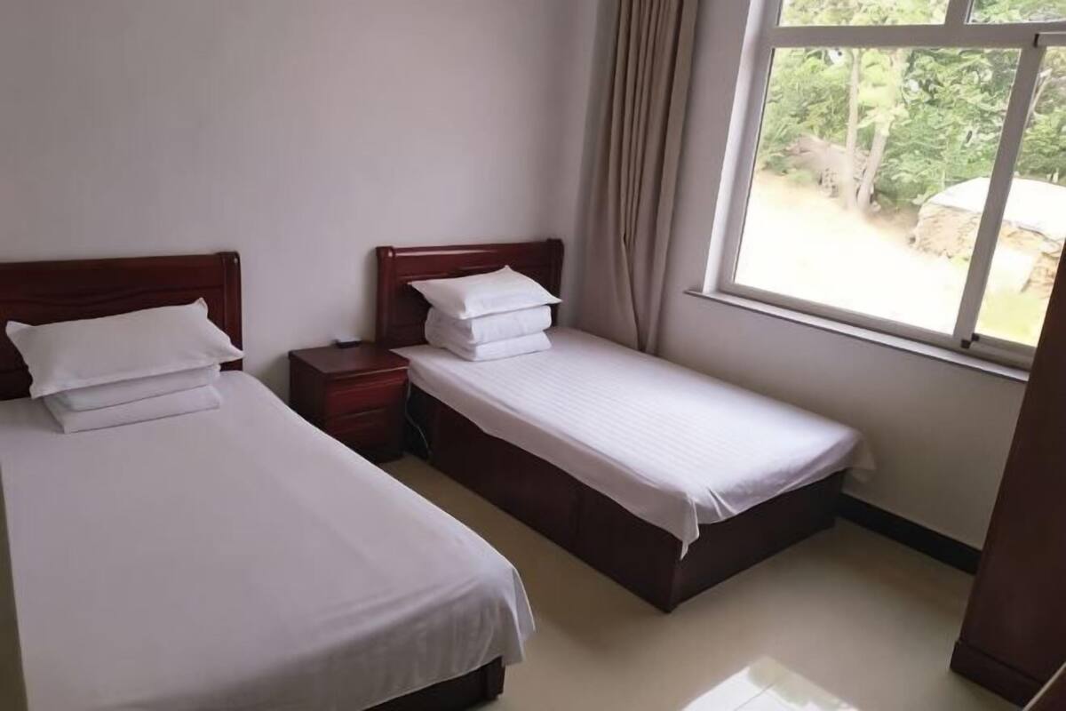 A room with two beds, a window with curtains, and a view of greenery outside.
