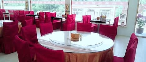 A dining area with round tables and red chairs, large windows with a view of greenery, and a white tablecloth on one table.
