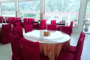 A dining area with round tables and red chairs, large windows with a view of greenery, and a white tablecloth on one table. - Bingyugou Jiahua Business Hotel (Zhuanghe)
