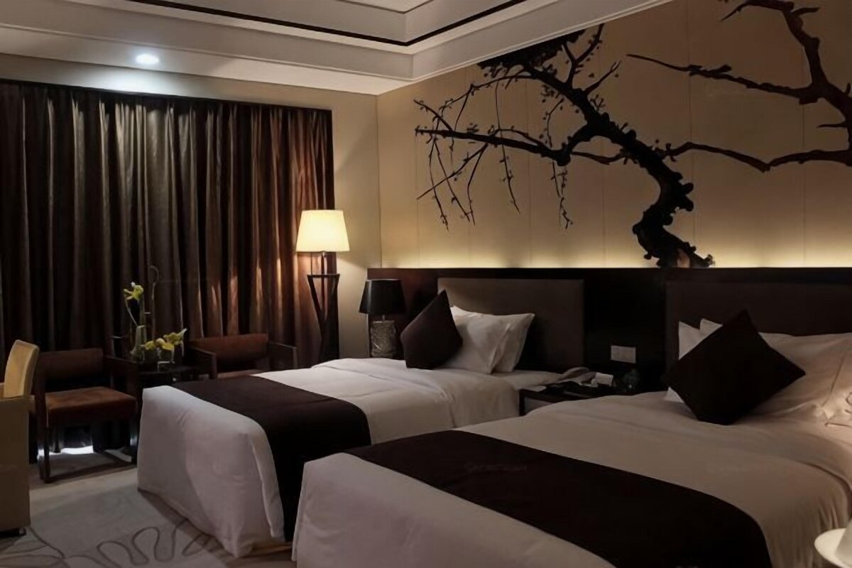 A hotel room with two beds, a desk, a chair, a lamp, and a wall-mounted artwork.