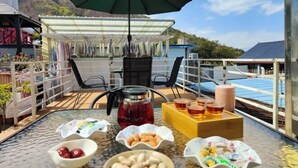 A dining area with a table set for two, featuring a variety of snacks and beverages, a green umbrella, and a view of the sea and hills in the background. - Xi Chang Blooming Azalea Hostel (Xichang)