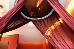 Una habitación con ventanal circular, cortinas rojas y un equipo de aire acondicionado a la vista.