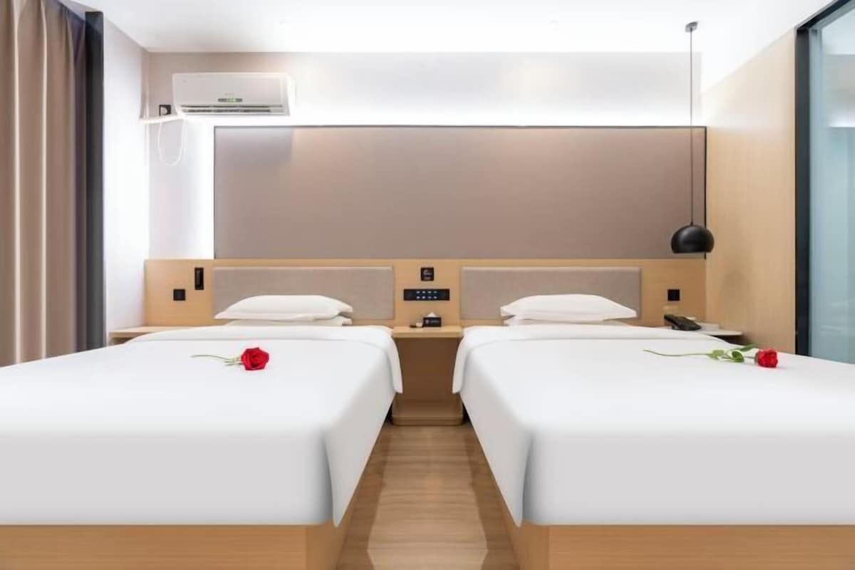 A hotel room with two beds, a wall-mounted air conditioner, a bedside table with a lamp, and a window with curtains.