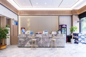 Zona de recepción de un hotel con mostrador de mármol, dos taburetes tipo bar, una máquina expendedora y una planta en maceta.