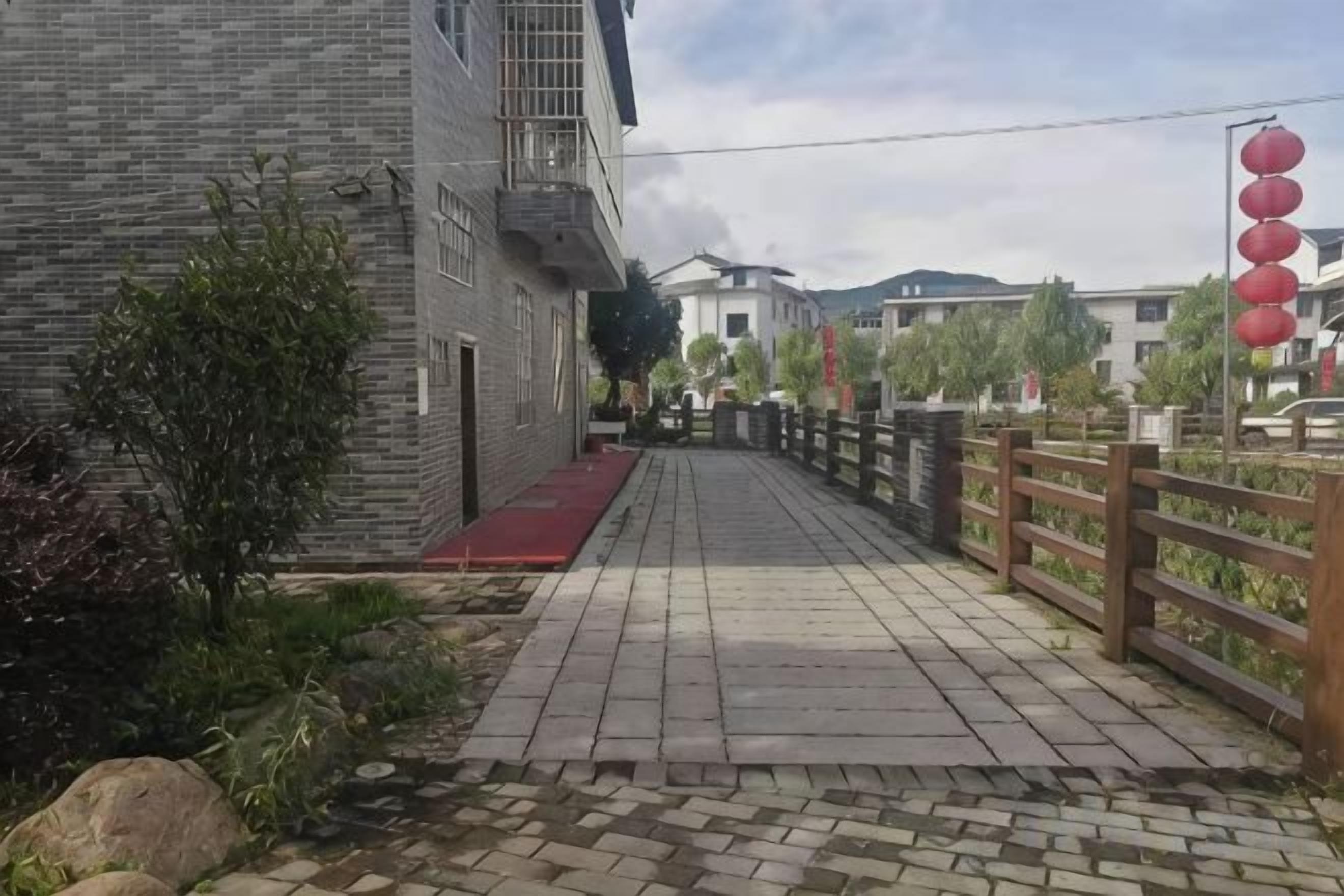 Un chemin pavé mène à un bâtiment surmonté d’une marquise rouge, avec une clôture en bois et des lanternes rouges disposées sur le côté droit.