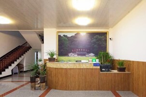 Un área de recepción con un mostrador de madera, plantas en macetas y un cuadro grande con la imagen de un edificio tradicional.