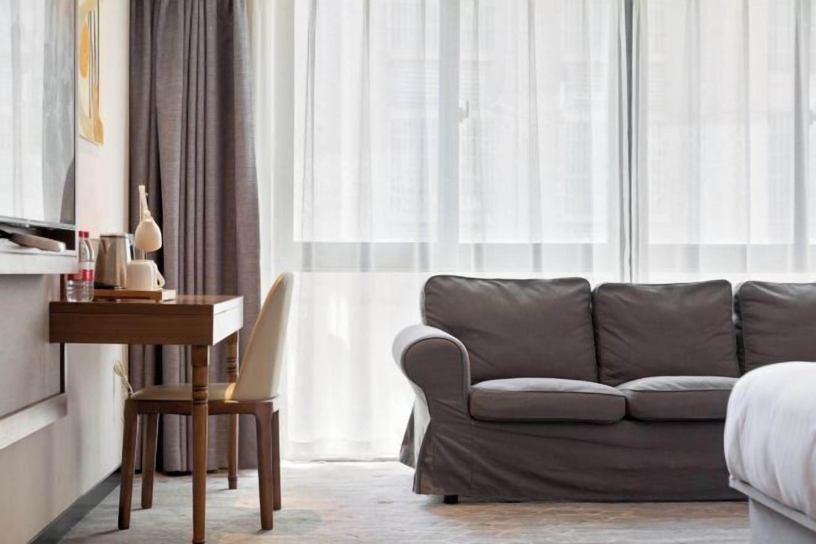 Quarto de hotel moderno com um sofá cinzento, uma mesa lateral de madeira com uma luminária e uma cadeira.