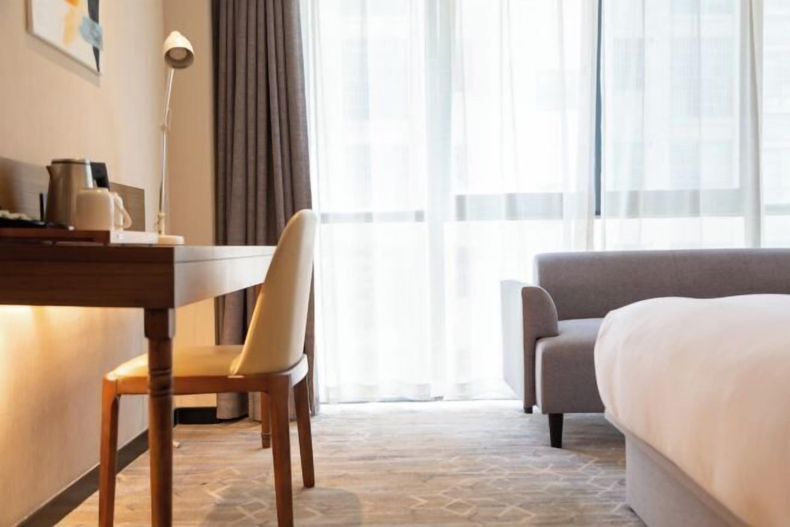 A hotel room with a bed, a desk with a chair, a lamp, and a window with curtains.