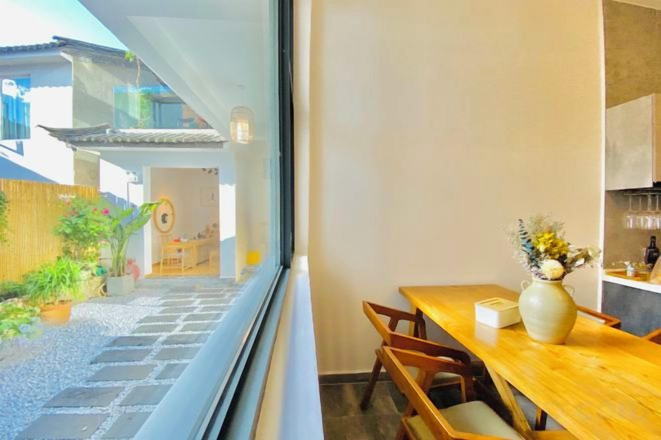 Un comedor con una mesa y sillas de madera, un florero con flores y vistas a un jardín y un edificio.
