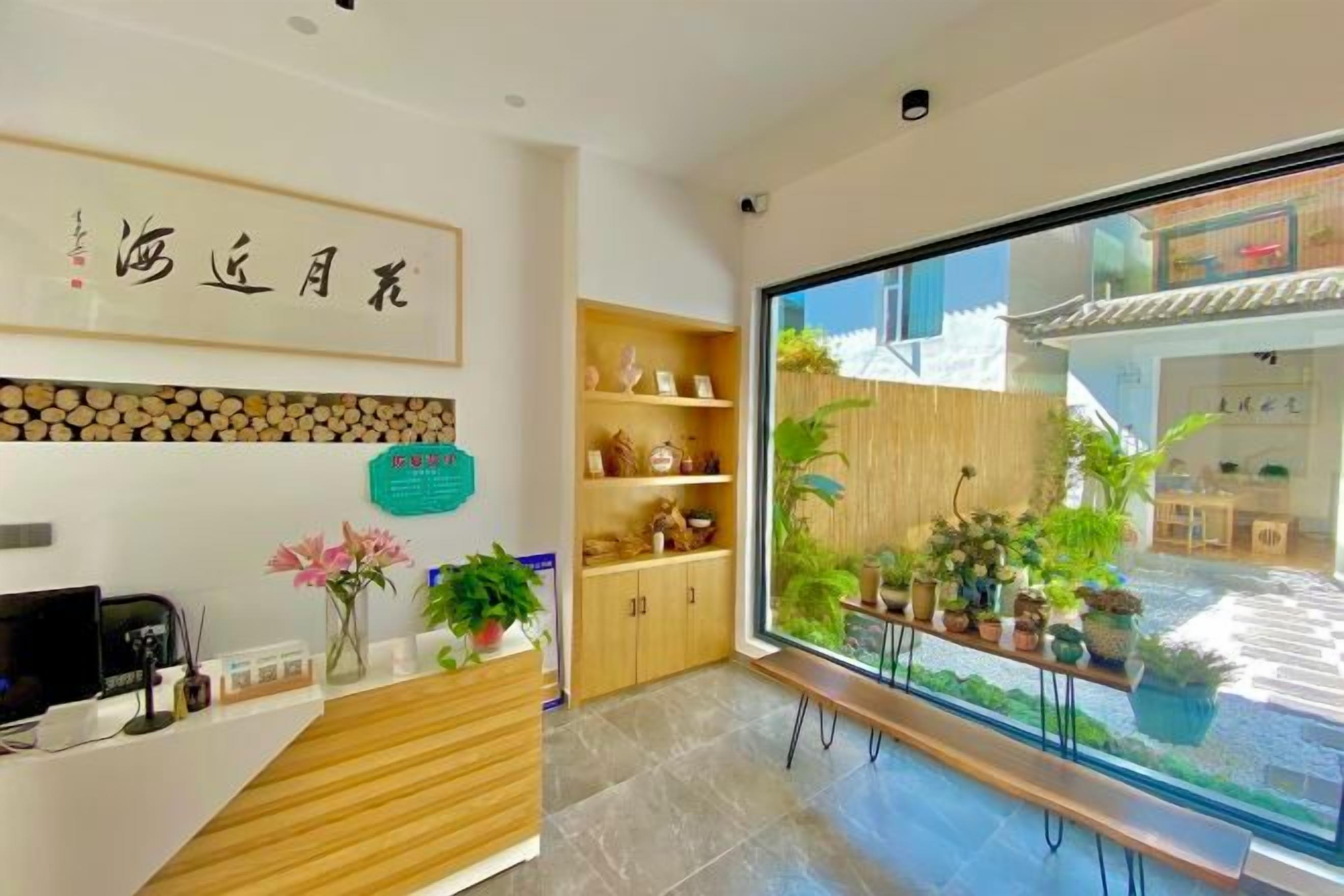 Zona de recepción con mostrador de madera, una computadora y un cartel. Hay estantes con objetos decorativos y un ventanal con vistas al exterior.